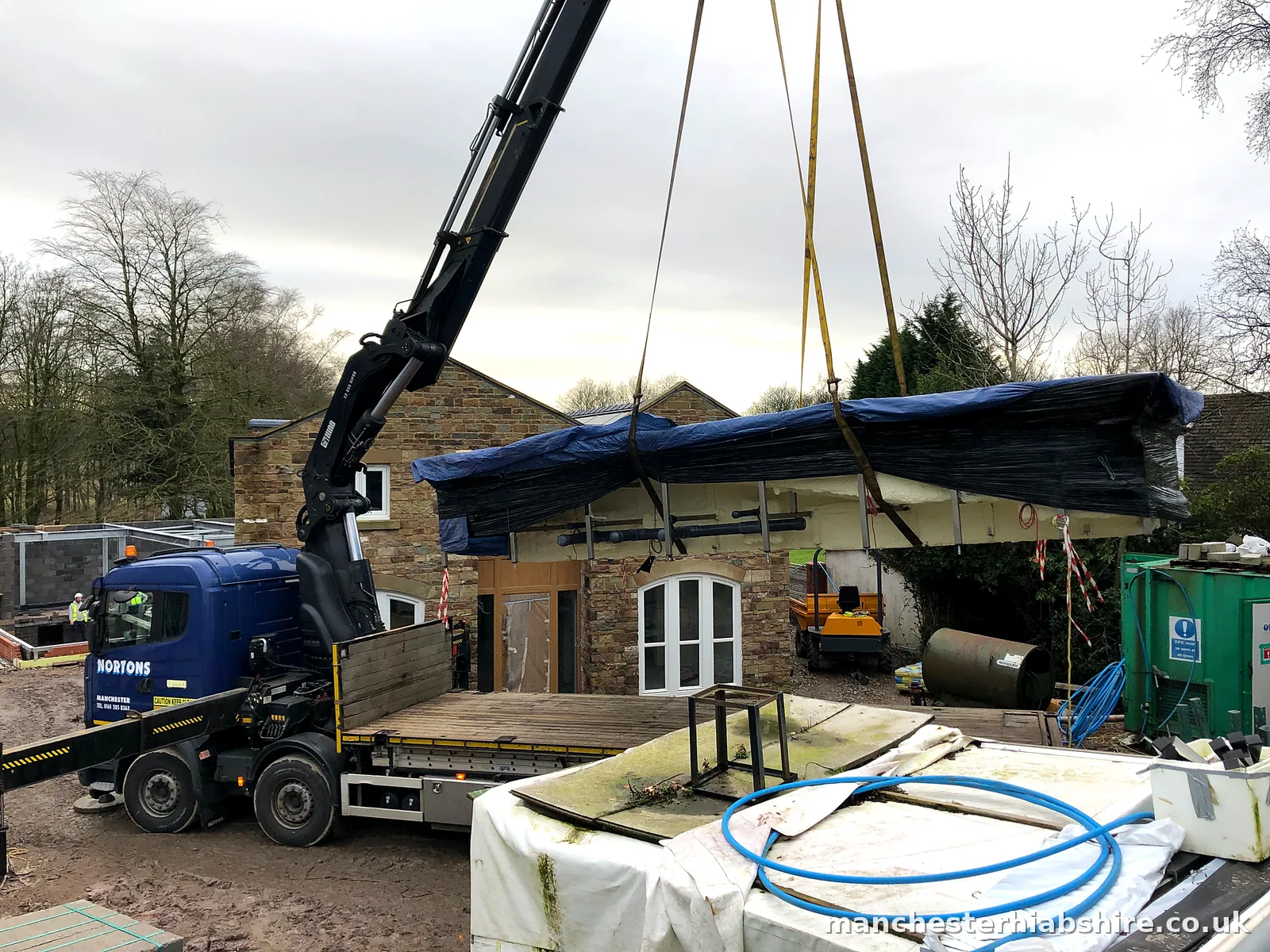 Long wrapped pool shell lifted over a stone building on the lorry mounted crane