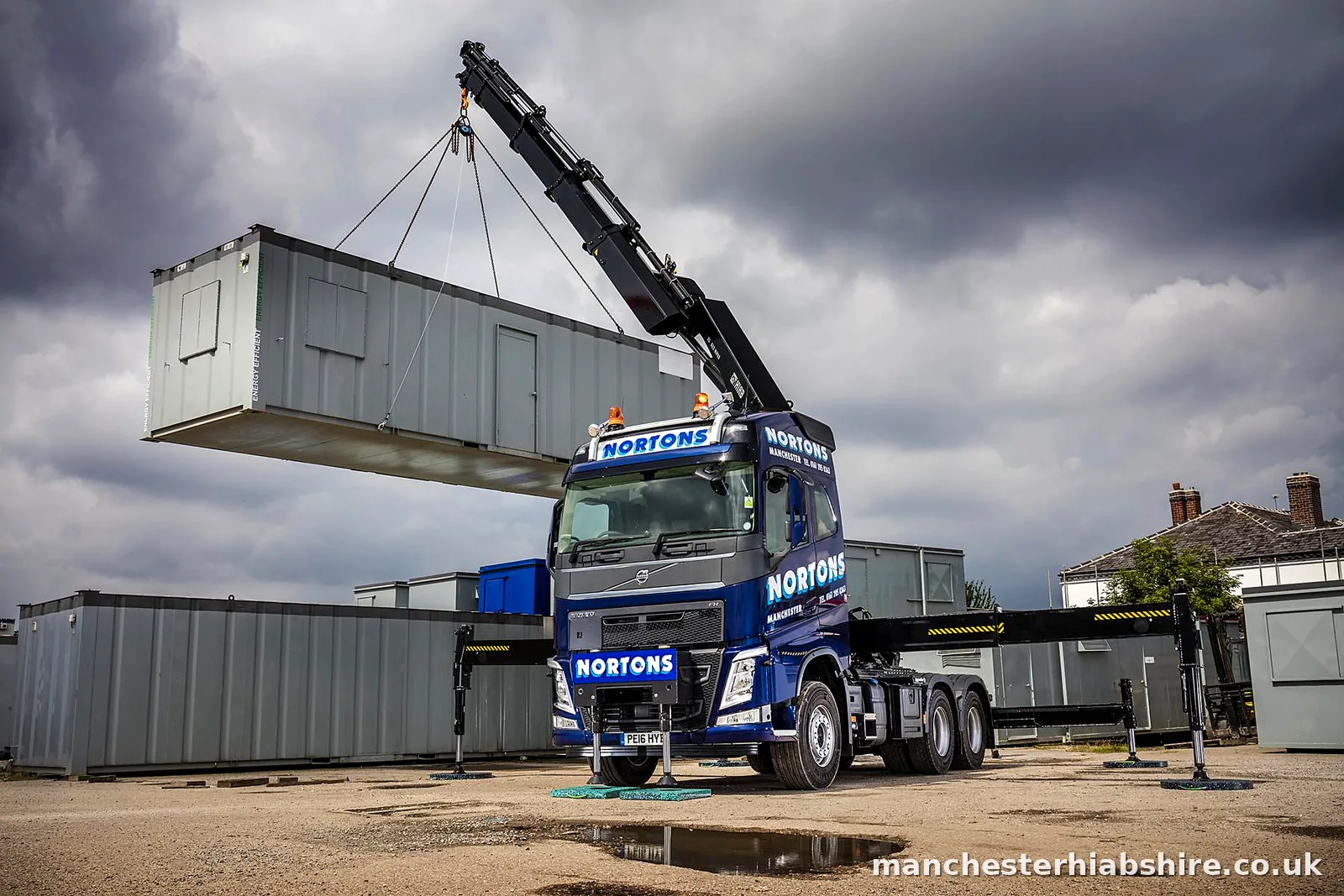 Lorry mounted crane holding a site cabin level under heavy cloud