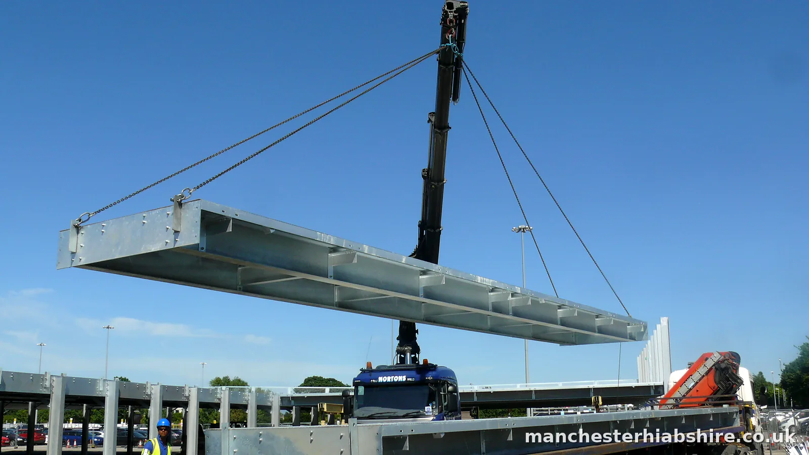Galvanised steel deck section lifted high against a clear sky with a banksman below