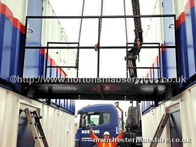 Banksman in full protective equipment guiding a steel walkway lift between cabin rows
