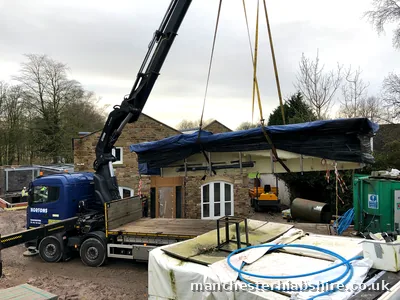 Long wrapped pool shell lifted over a stone building on the lorry mounted crane