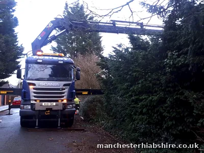 Lorry on a narrow residential road with outriggers down and the boom reaching over a tall hedge