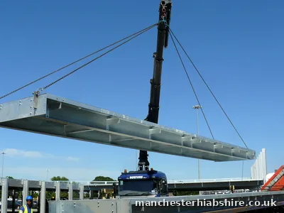 Galvanised steel deck section lifted high against a clear sky with a banksman below